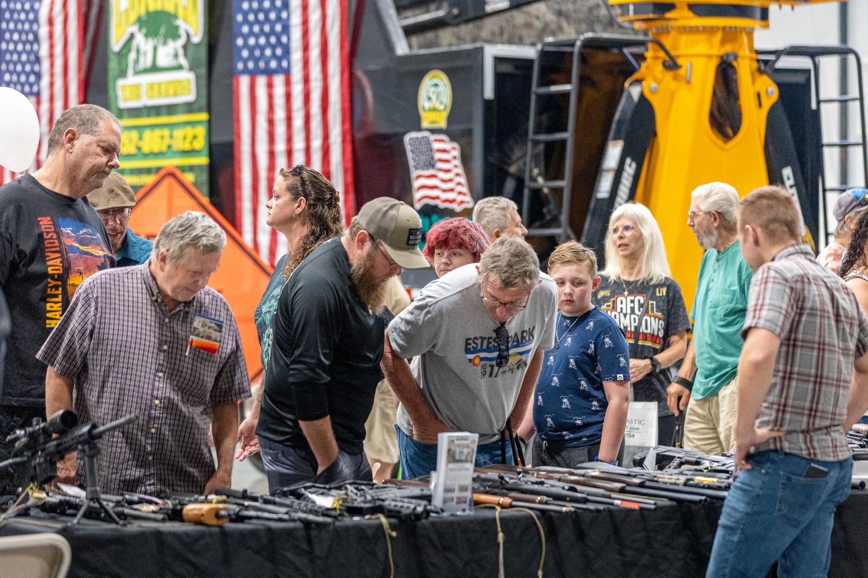 Several men browse a table filled with a variety of firearms and weaponry for hunters. American flags and large environmental equipment are in the background