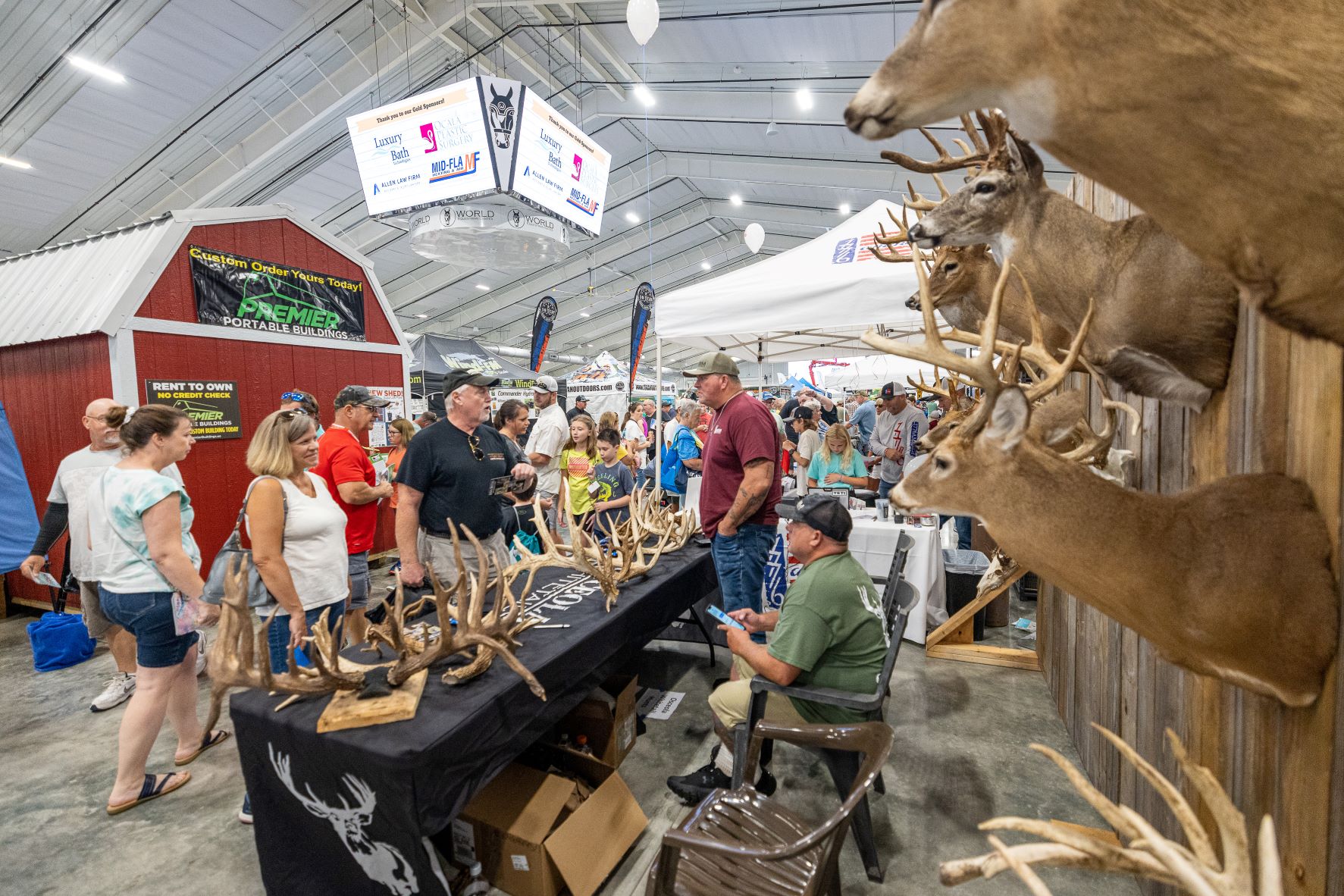 A display booth showcases several mounted deer with antlers on the table. A seated man talks happily with several onlookers.