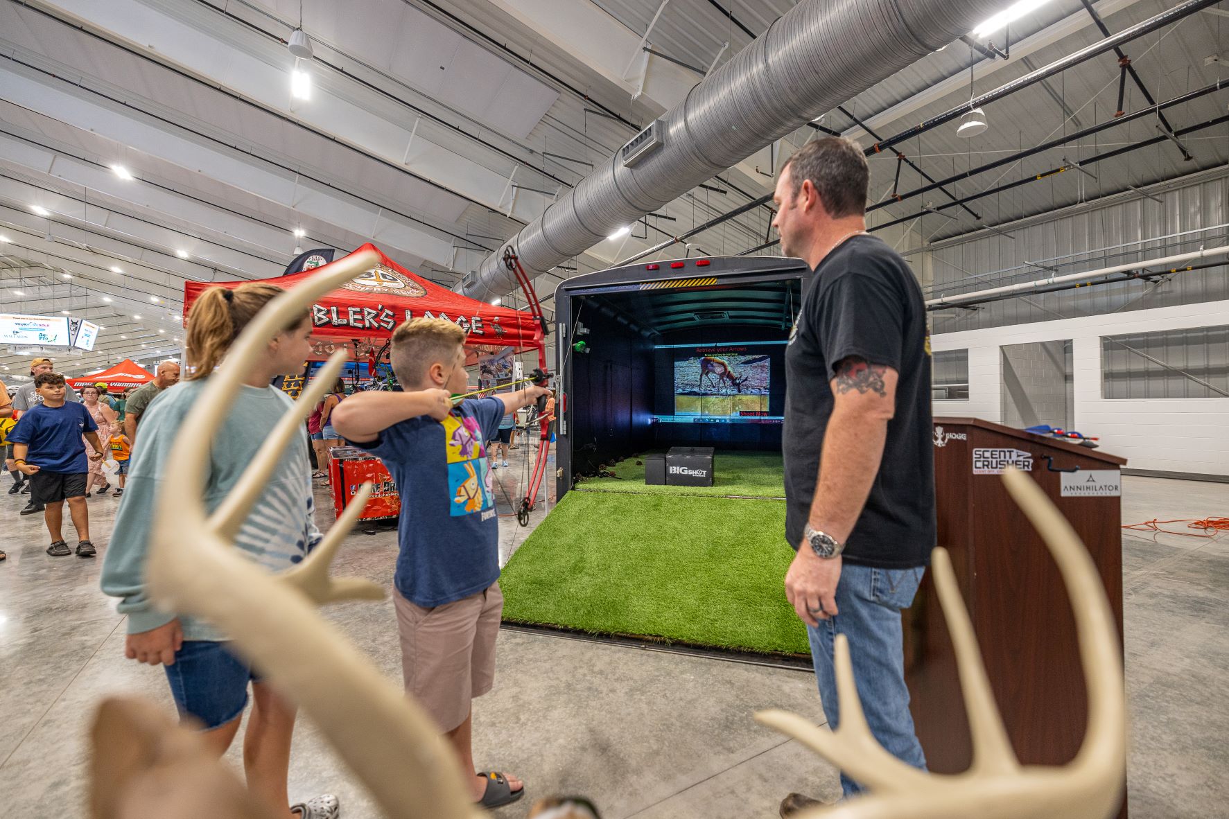 Small child practices 3-D archery at a demonstration set up in a trailer. An older girl watches over his shoulder, smiling. 